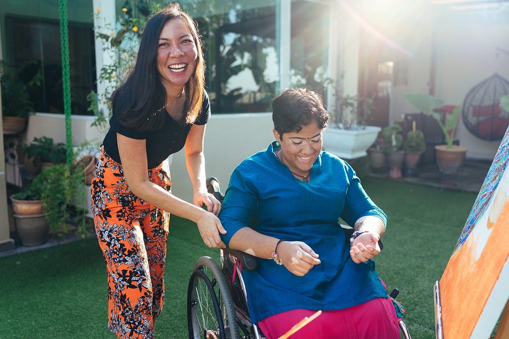 Rozella smiling and pushing artist, Aveena Devi in a wheelchair in the garden 
 under a soft daylight