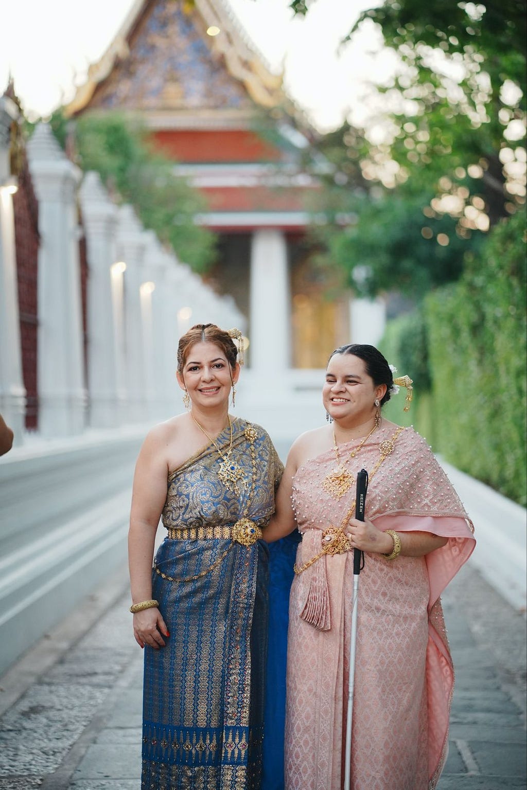Dos mujeres están de pie y portan vestimenta tradicional tailandesa mientras comparten un abrazo mirando hacia la cámara. Una de ellas usa un bastón.