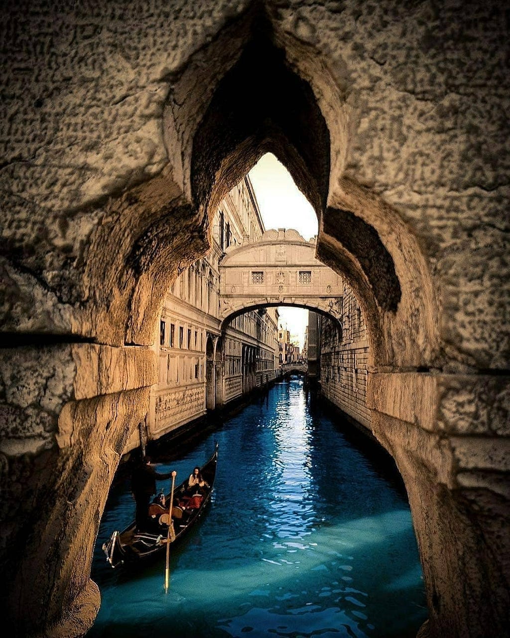 The view through an archway in one of the canals in Venice. The stone archway is old and weathered. The water is a deep navy blue; there is a boat with a couple in the lower foreground being rowed in a small boat.