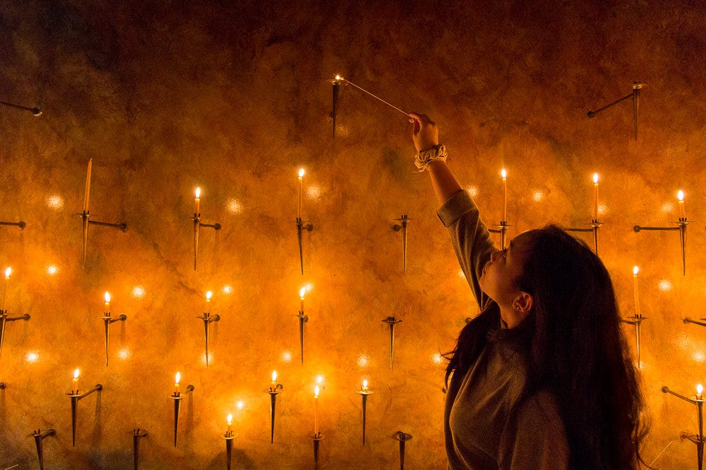 A young girl is lighting a prayer candle on a wall. The flames make the whole photo illuminated with an orange glow.