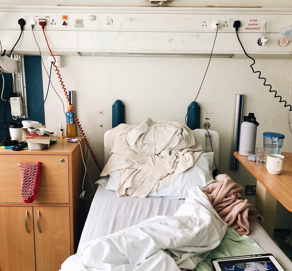 a hospital room with an unmade bed and blankets strewn across it. different drinks scattered around the two bedside tables. an ipad sits on the end of the bed, and a book is propped on the bedside table on the left. wires hang down across the back wall.
