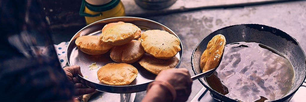 A person is making pooris (a deep-fried bread common in South Asian cuisine) lifts one from the oil. In their other hand there is a plate containing the rest of the batch.