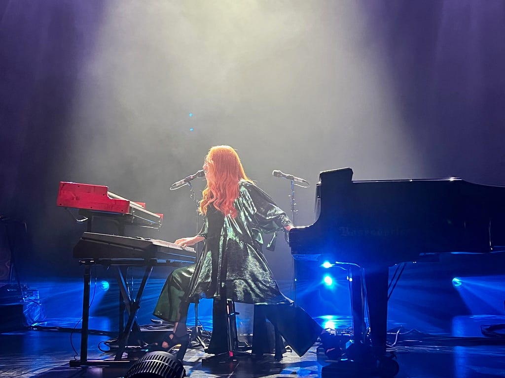 An image of singer-songwriter Tori Amos onstage in Seattle, wearing a green spangled outfit while playing her Bösendorfer piano with her left hand as her right plays a synthesizer.