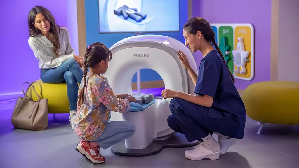 An image of the MRI scanner designed for pediatric patients. A little girl places her teddy bear into the scanner while a nurse assists her. On the left, another woman — likely the mother — observes the scene.