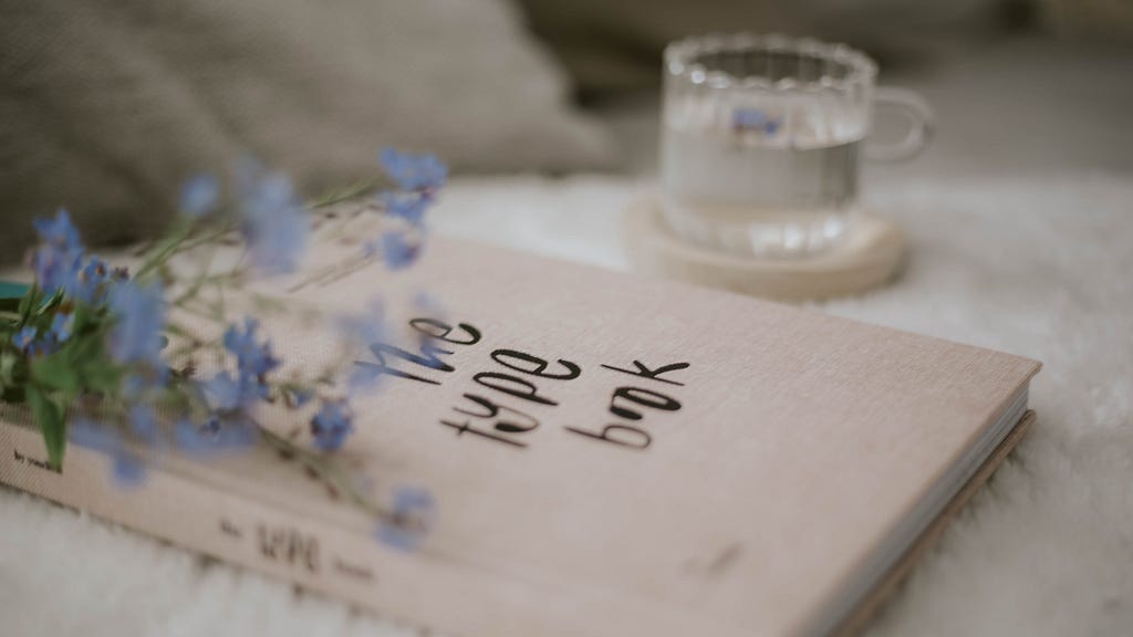 A visually pleasing image of a book titled “The type book”, a branch of delicate blue flowers and a glass of water. All presented in a soothing and romantic pastel color scheme.