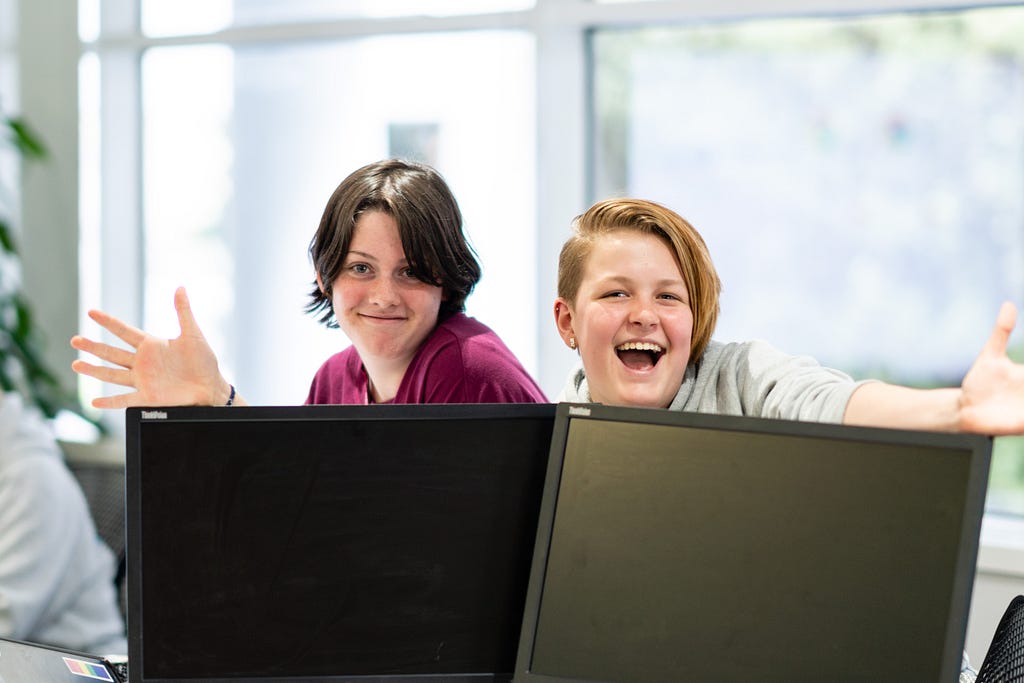 Two young people with their hands out smiling behind two laptop monitors