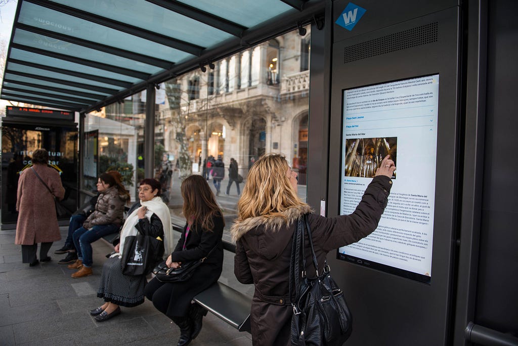 A woman in a bus shelter in Barcelona, Spain, reads a video screen for information.