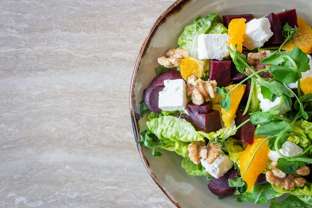 A bowl of salad with fruit, greens, beets and cheese on a gray table