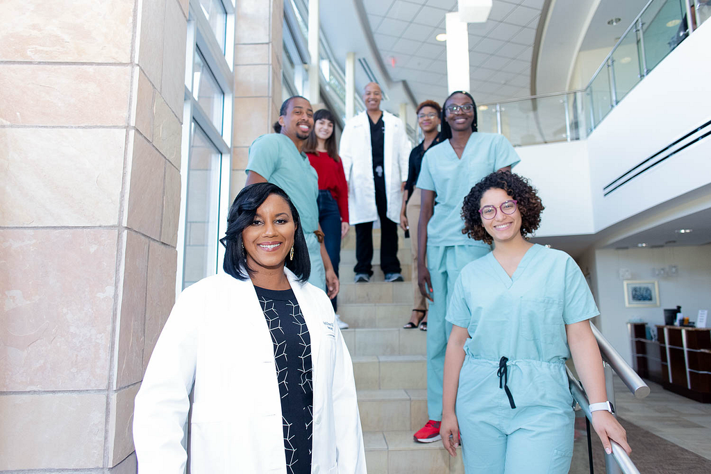 A diverse group of medical professionals and students pose on a stairwell inside a modern building. Dr. Porter is standing in the front wearing a white medical coat. Around her are students wearing scrubs and casual attire, all smiling and looking engaged. The setting has large windows and bright lighting.