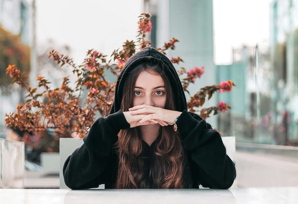 Sadly reflective young woman in a dark hoodie sitting with her elbows on the table, hands clasped in front of her mouth.