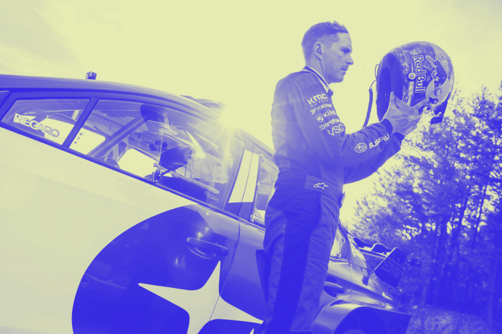 Scott Speed wearing a blue race suit in front of a blue Subaru, frowning at his helmet about to put it on.