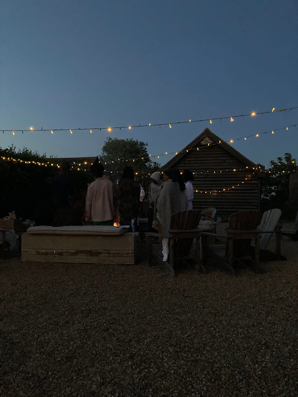 A dark night time shot of guests around a fire pit. The fire is low. Guests wear blankets around their shoulders. There are strings of fairy lights over head.