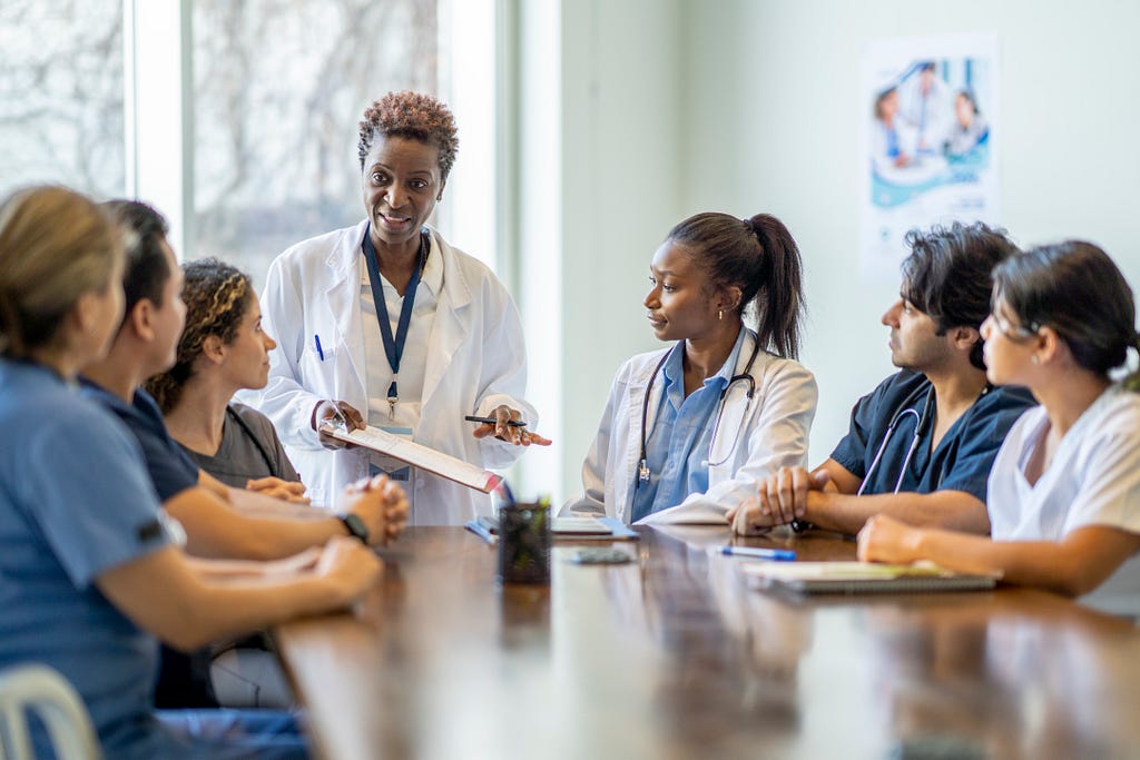 A diverse group of health care workers at a conference table listening to a Black doctor. Photo by FatCamera/Getty Images