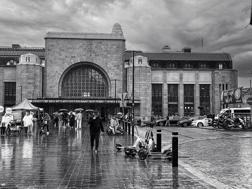A black and white image of the Helsinki Railway station with scooters and cars parked nearby and people walking on wet cobblestones under umbrellas.