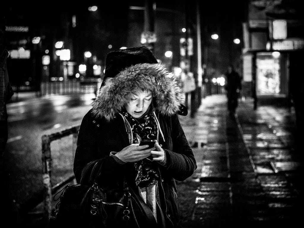 on a rainy night at what looks like a bus stop, a woman in a thick winter coat looks at a smartphone, the screen illuminating her face and making it uncharaceristically pale