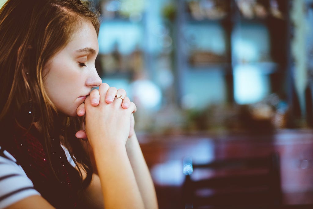 A woman is in a church, her eyes closed and hands clasped in front of her, in a posture of prayer.