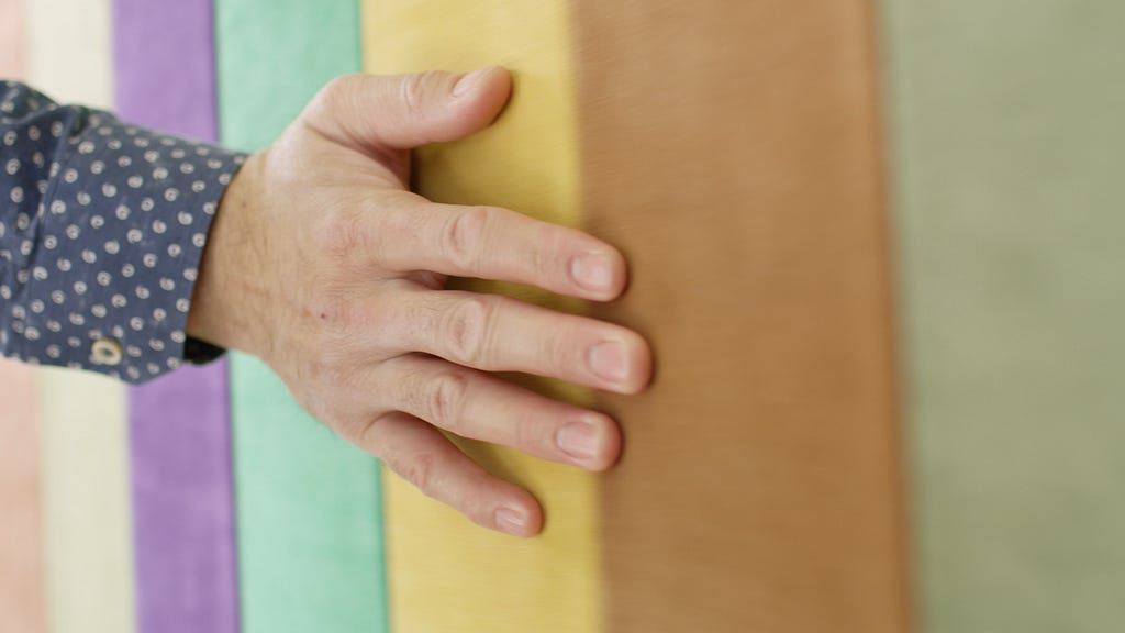 Close up of a man’s hand, open palm, runs along pastel purple, green, yellow and brown colored Alcantara textile.