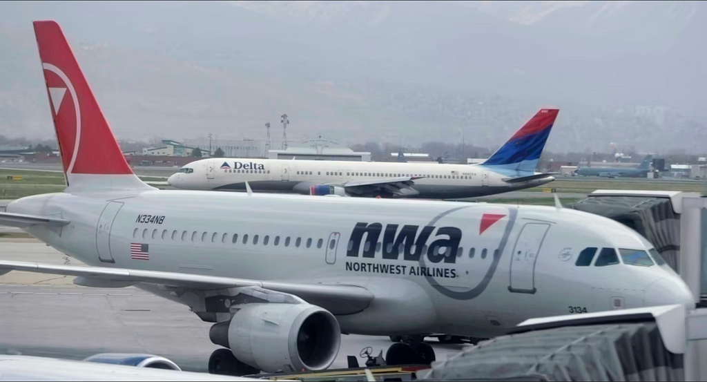 A photo of two planes. In the foreground is a Northwest Airlines Airbus A319, with the Northwest Airlines silver livery. It has a silver fuselage, a red tail, and “nwa” in large letters on the side of the plane. In the background is a Delta Airlines 757, with the Delta Airlines “Colors In Motion” livery. It has a white fuselage and a tail painted to look like a red, blue, and light blue flag. The side of the plane says “Delta.”