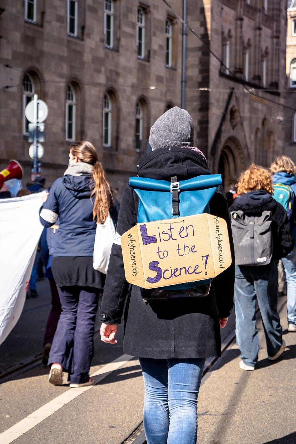 People marching with a poster saying “Listen to the Science!” on someone’s backpack