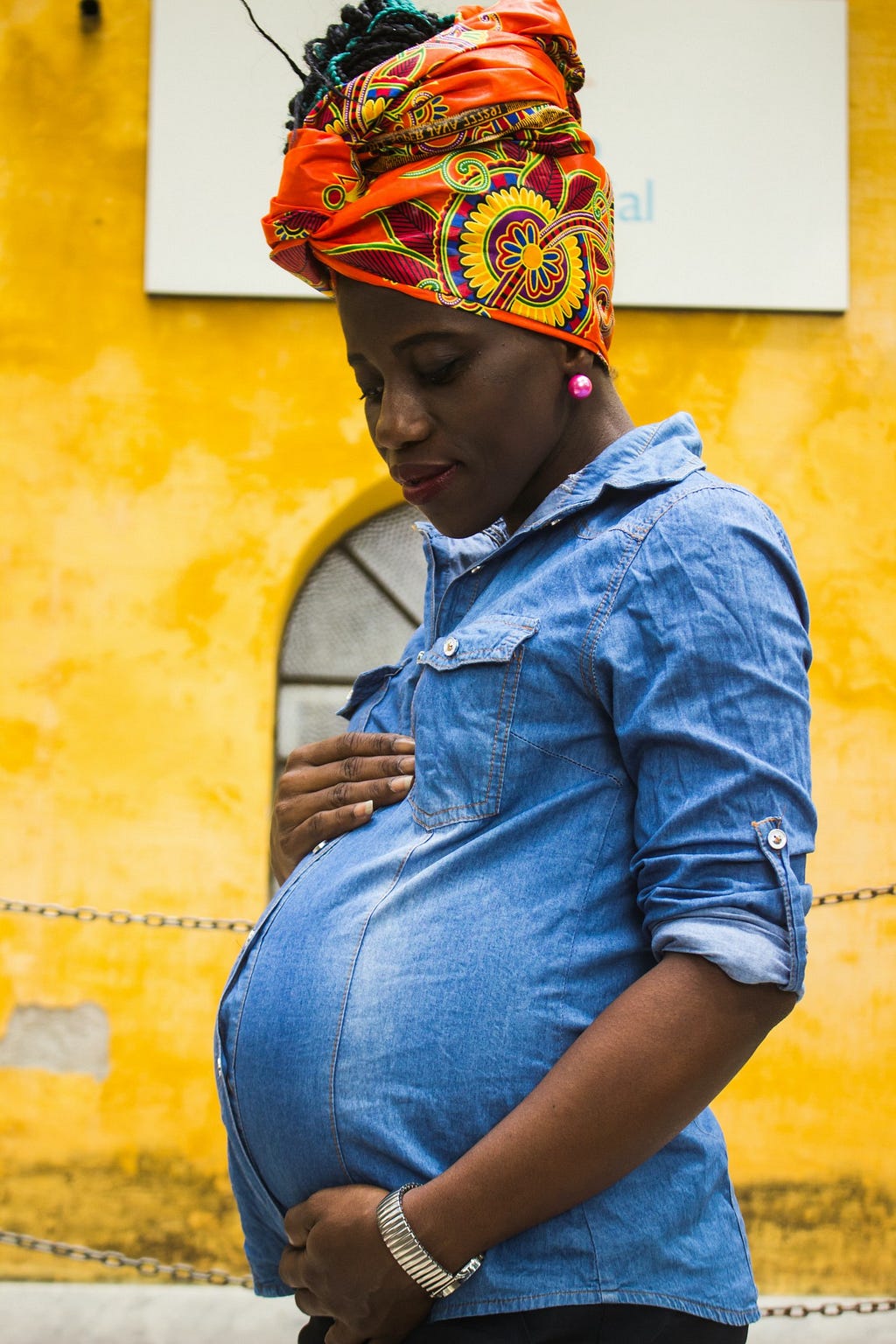 a pregnant femme person with dark brown skin, holding her belly. She wears a blue-jean t-shirt. She has a brightly colored orange, yellow, and green headwrap over a braided updo hairstyle.