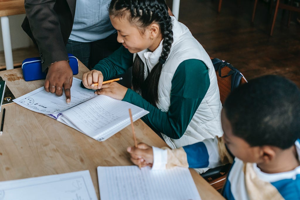 Two school children sit at their desk while a teacher helps one with their assignment.