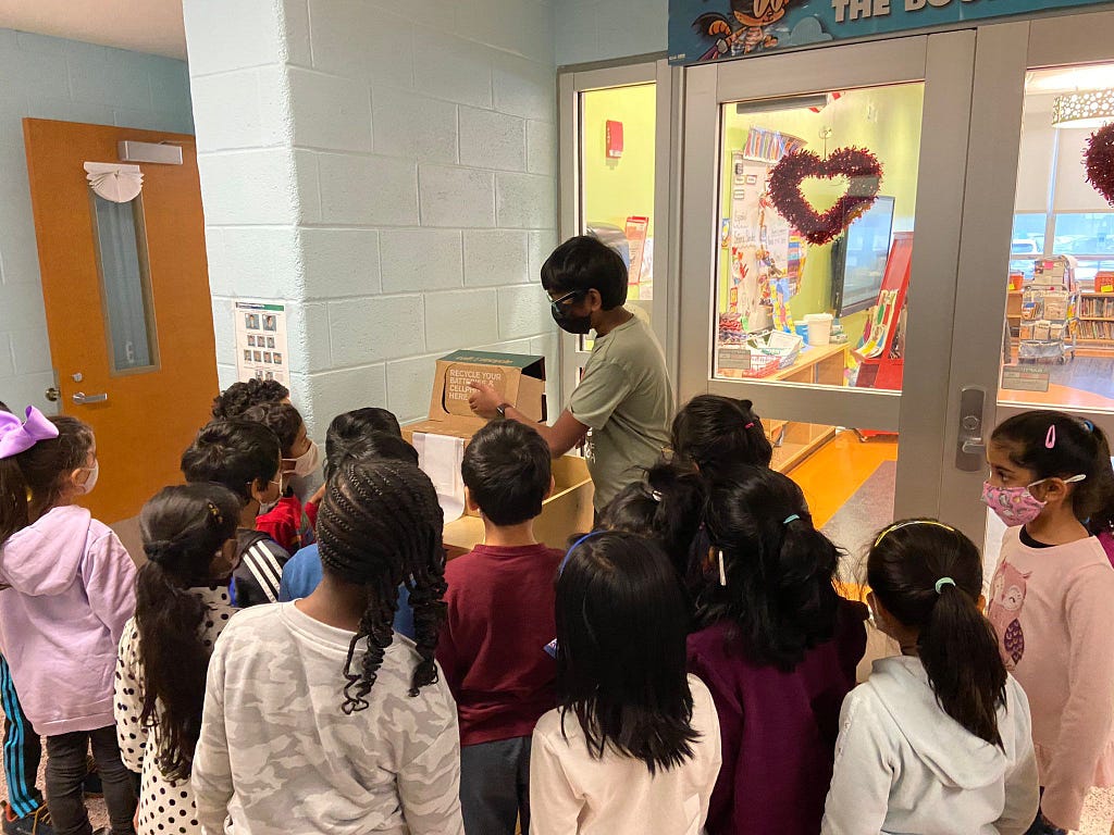 Same boy in mask demonstrating recycling on a trash can to a group of elementary schoolers. Everyone is wearing masks and all the kids have their faces turned away from the camera. There is a Valentines Day heart on the school door behind them.