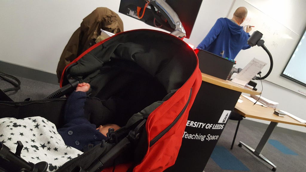 An image of Emily’s baby son sleeping in his pram in a classroom on the University of Leeds campus, with a member of LLC teaching staff in the background delivering a session on the Child and Family Studies degree.