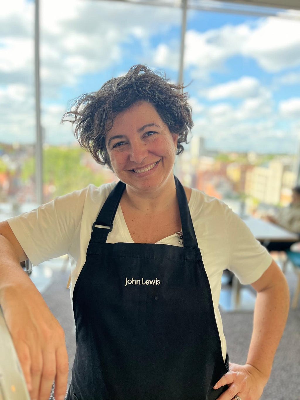 A picture of a smiley Chiara, wearing a white t-shirt and a black apron with the John Lewis brand on it.