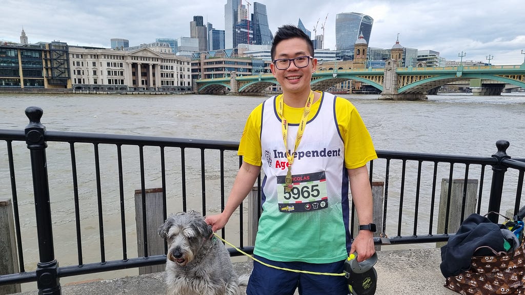 Nicolas, a man with short dark hair and glasses, smiles at the camera as he holds onto Elia, his dog who has grey and white scruffy fur. Nicolas wears his Independent Age running vest and a running medal.