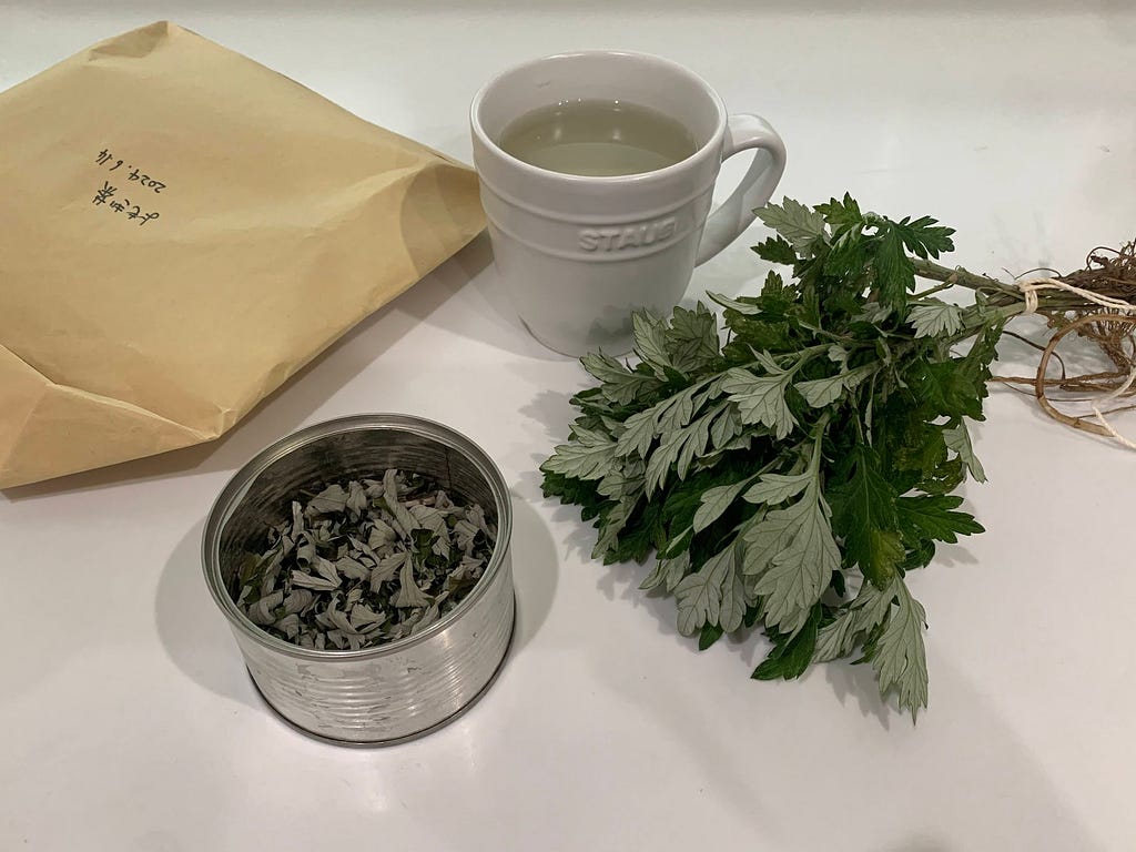 A setup for making homemade yomogi tea, featuring a bunch of fresh yomogi leaves, a container of dried yomogi, a mug of hot water, and an envelope for storing dried leaves.