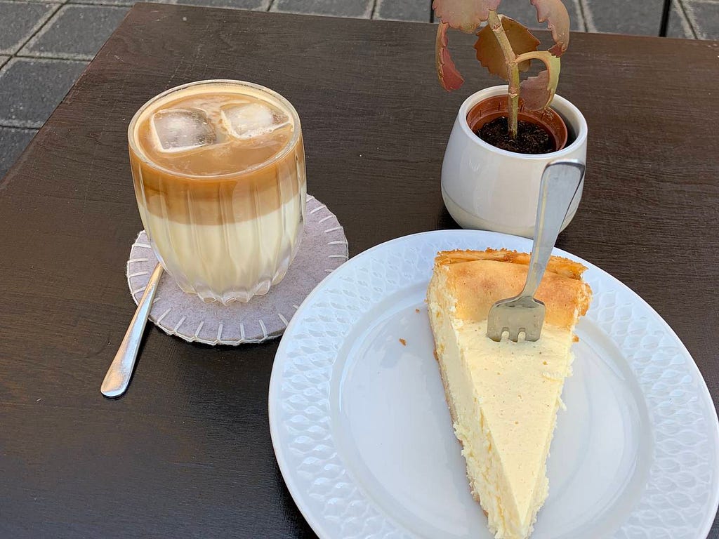 A coffee with milk and ice cubes, and a piece of cheese cake on a white plate