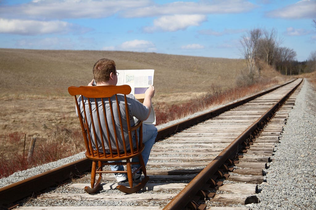 Man sitting in a chair casually reading a newspaper in the middle of a railroad track.