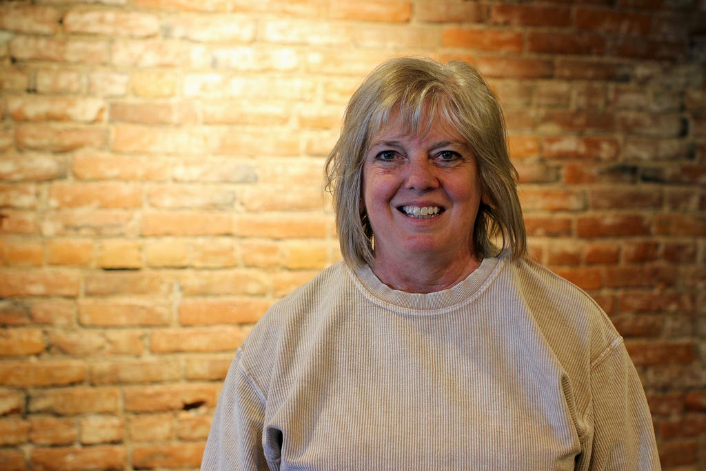 A woman poses in front of a brick wall brightened by an overhead light and smiles at the camera. She has shoulder length blonde hair and is wearing a cream colored sweater.