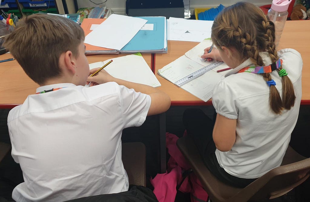 A photograph of two children wearing Design Club lanyards, sat together at a school desk sketching and writing in app design workbooks.
