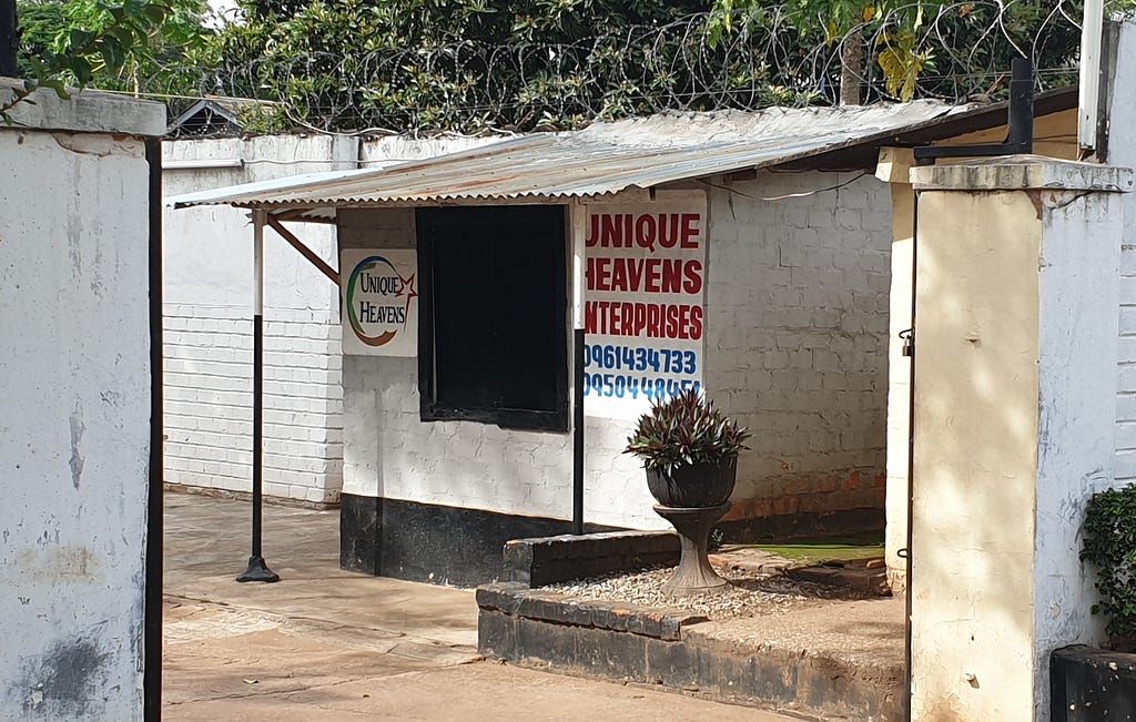 Little shop with “Unique Heavens Enterprise” sign on white walls, under a tin roof, seen through a gate.
