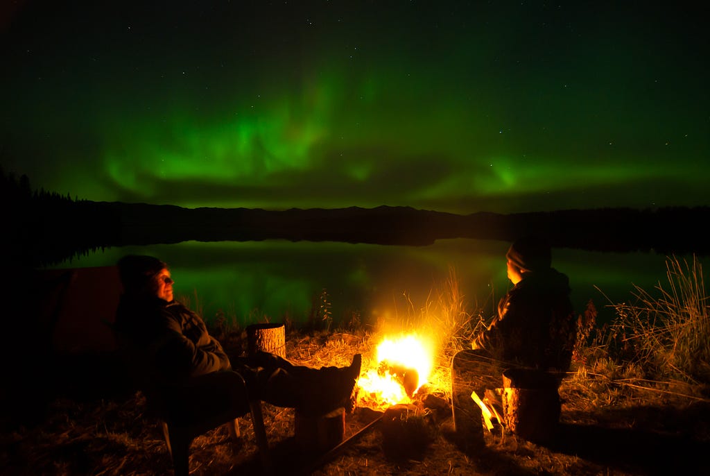 Two men sit by a fire near a river with green bursts of northern lights on the horizon.