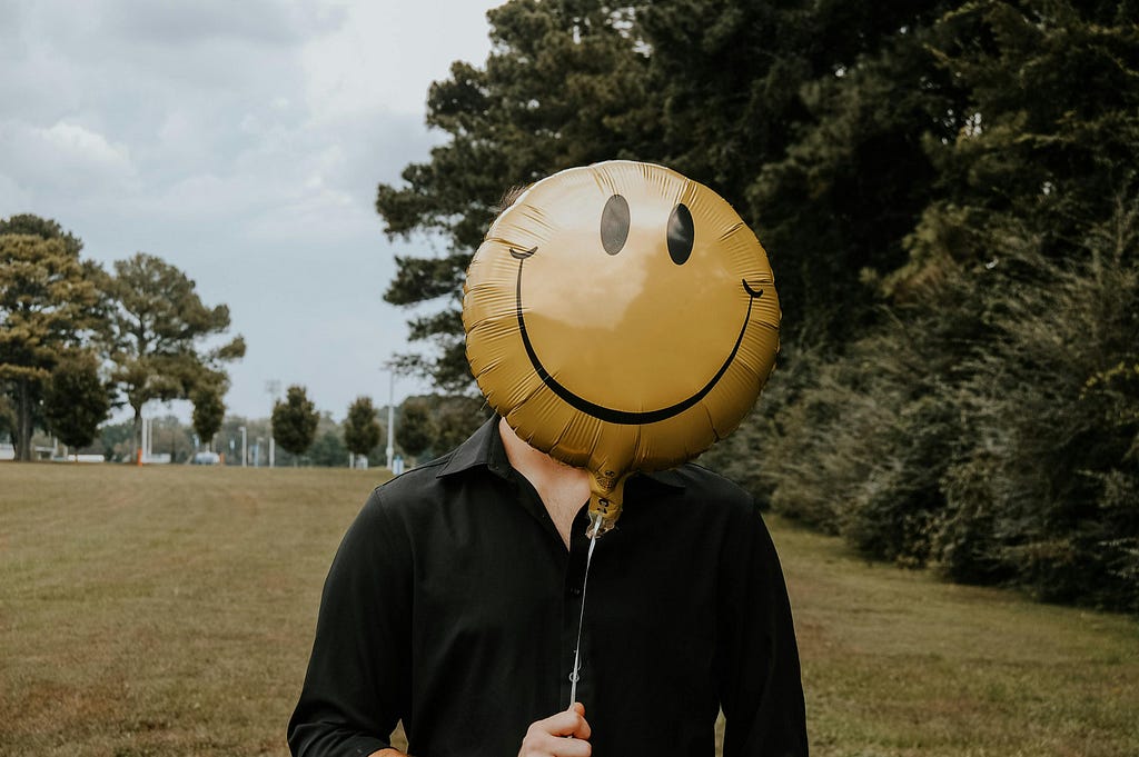 A person in a black shirt standing in a park, hiding their face behind a smily-face balloon.