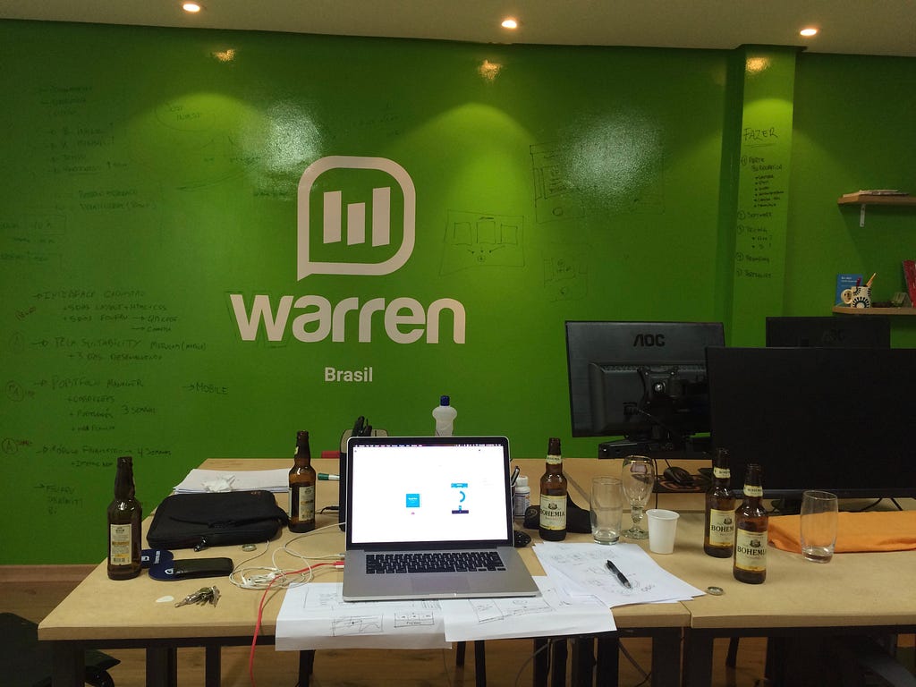 Foto de um notebook em cima de uma mesa de trabalho com folhas cheias de rascunho e garrafas de cerveja vazias. Ao fundo, uma parede verde com a primeira versão da logo da Warren.