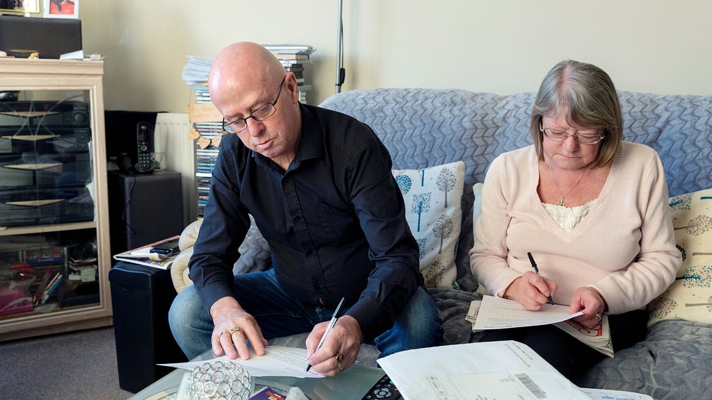 Photo of a man and woman sat on a blue sofa whilst filling out paperwork.