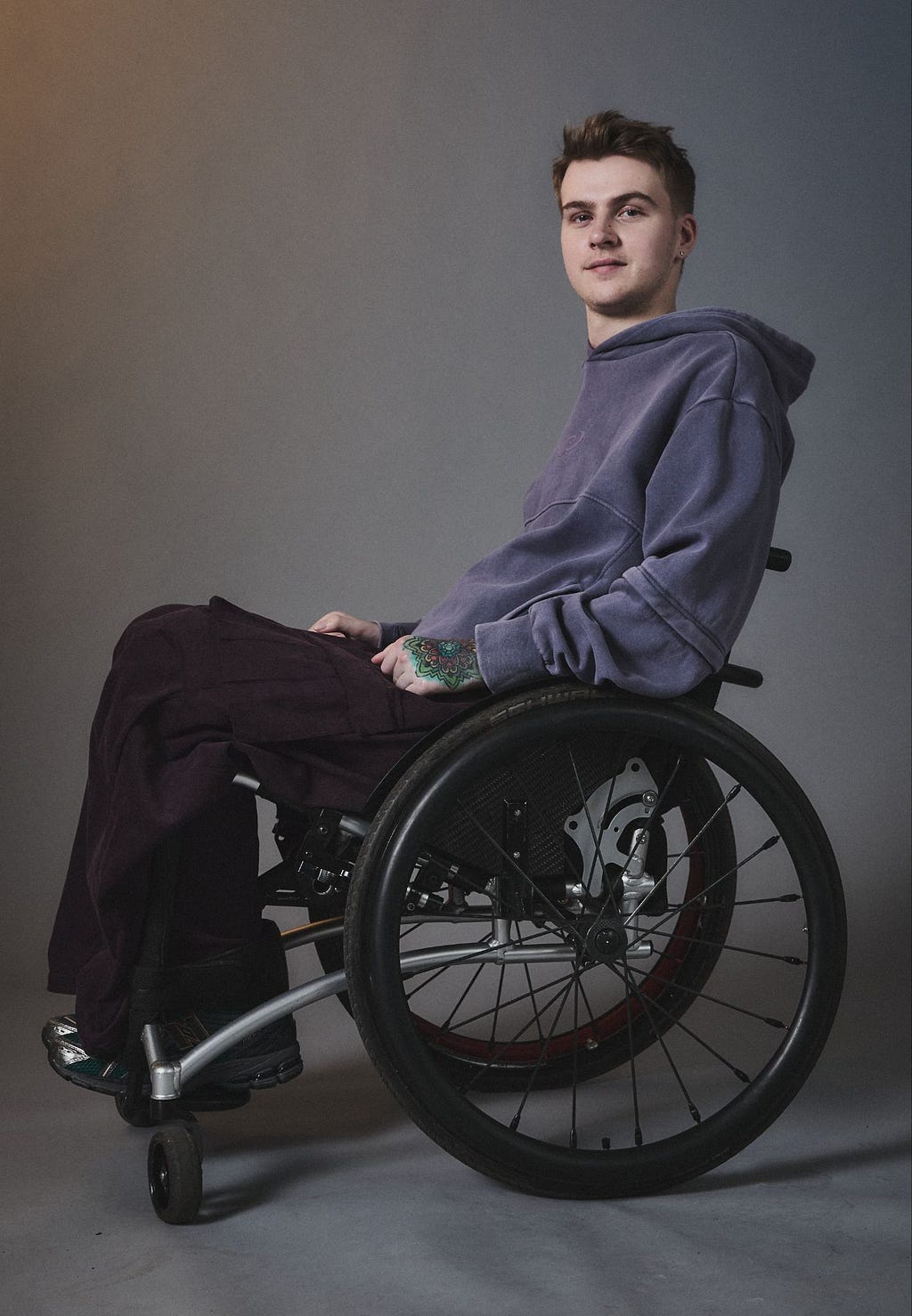 A white person with short brown hair wearing a muted dark purple hoodie and black trousers, using a wheelchair and pictured against a grey gradient background.
