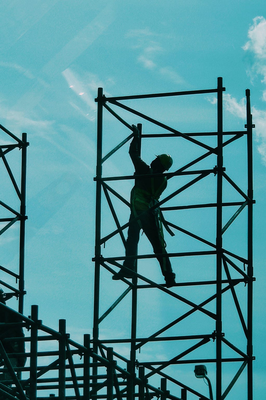 A construction worker stands precariously on scaffolding, suspended in the air, under a blue sky.