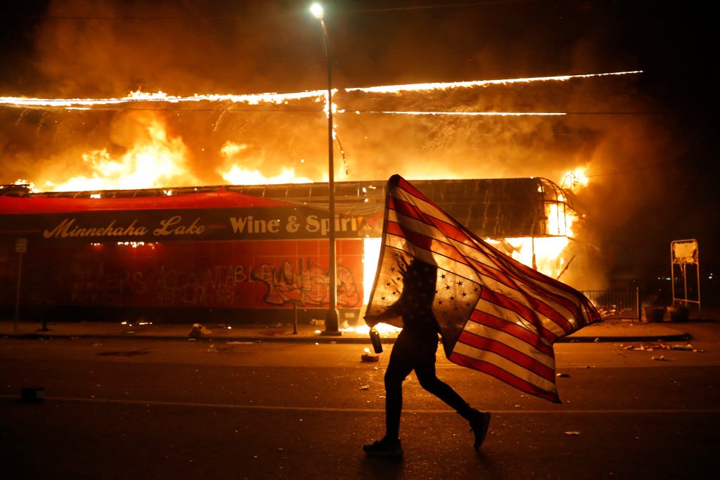 A silhouetted person shrouded by an American flag walks past a burning corner store.