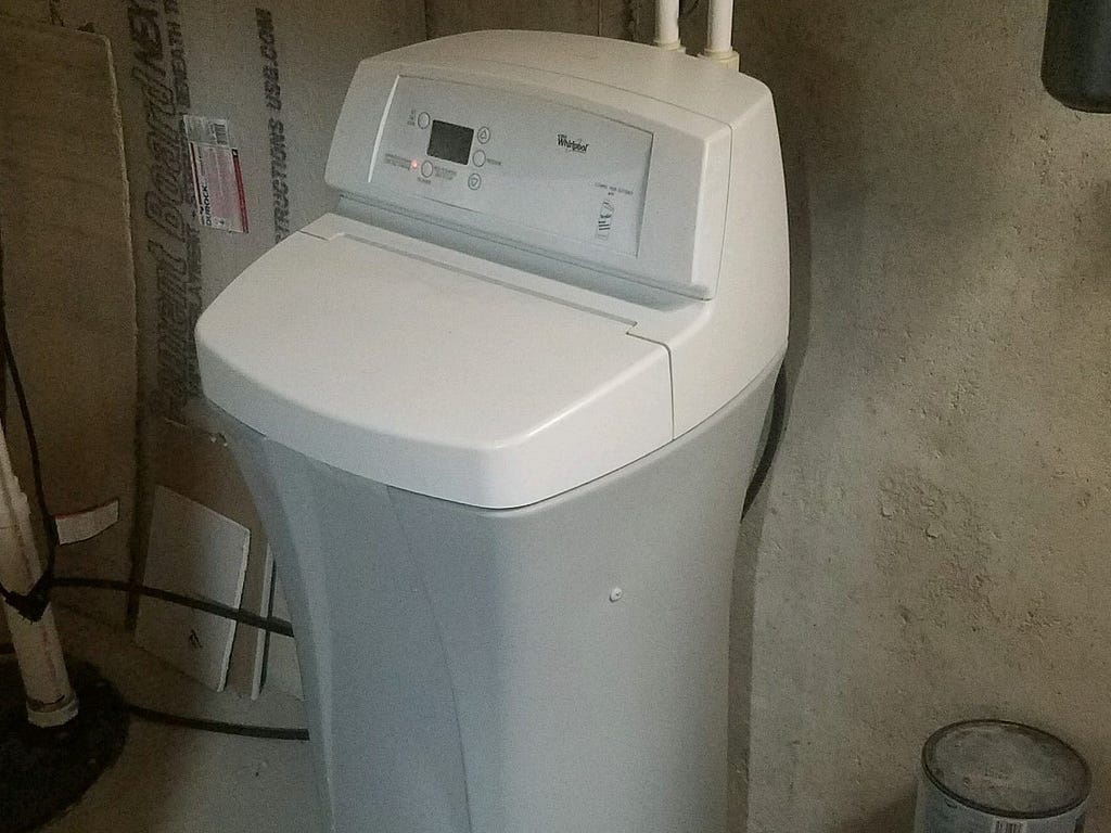 A photo of the author’s water softener. It has a white top with control buttons and display, and a grey colored bin for the salt.