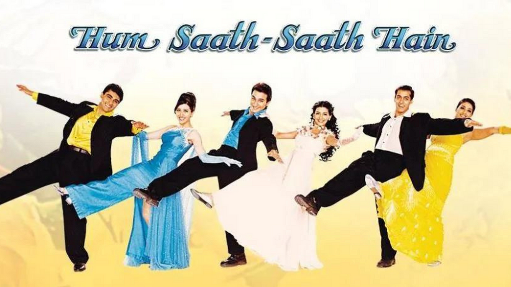 A poster shows 6 actors — 3 men and 3 women — and the words Hum Saath Saath Hai is visible on the top in blue. The 6 actors stand in a straight line with one leg in the air and both arms raised up on either side. The men are in black suits. The women are in traditional Indian attire and in gowns.