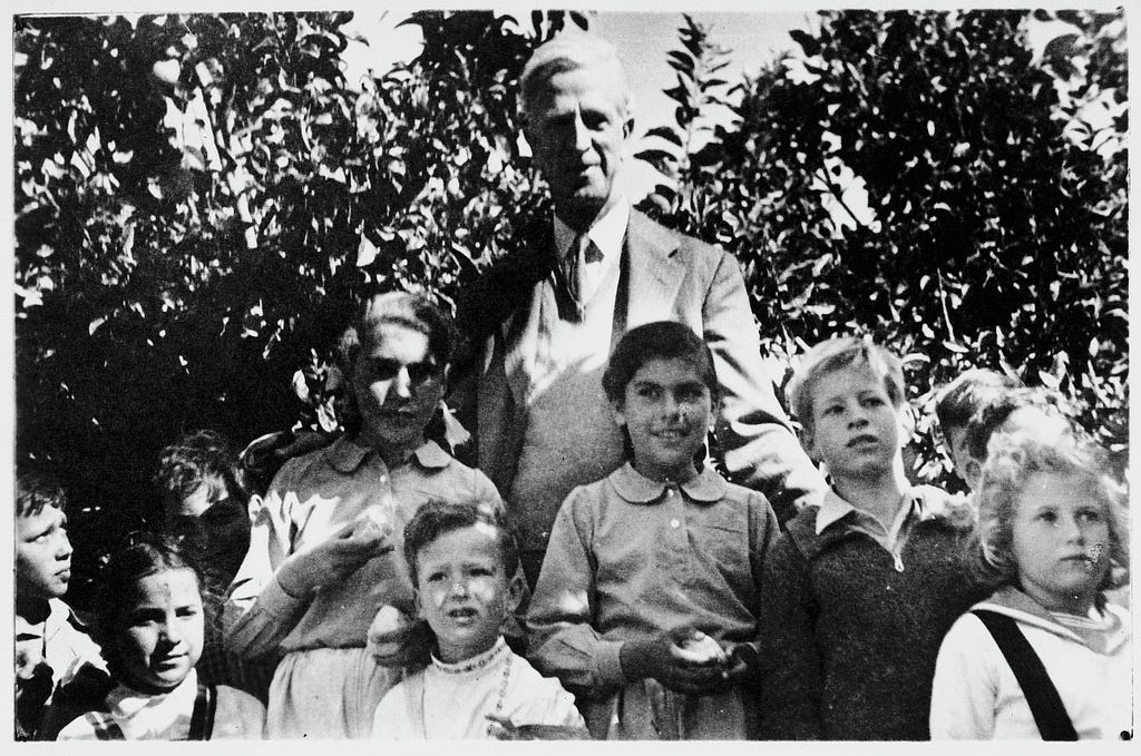This photograph features the same man, with notably aged features. He wears a sport coat, sweater, tie, and shirt. he is standing with a group of about ten children. They are in dappled sunshine in front of trees.
