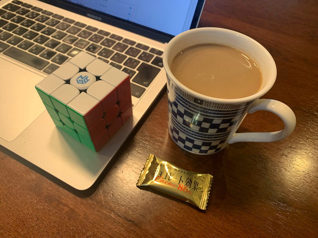 A laptop with a Rubik’s cube on top of it, next to a mug of coffee and a piece of chocolate on a wooden table. The scene depicts a morning routine setup.