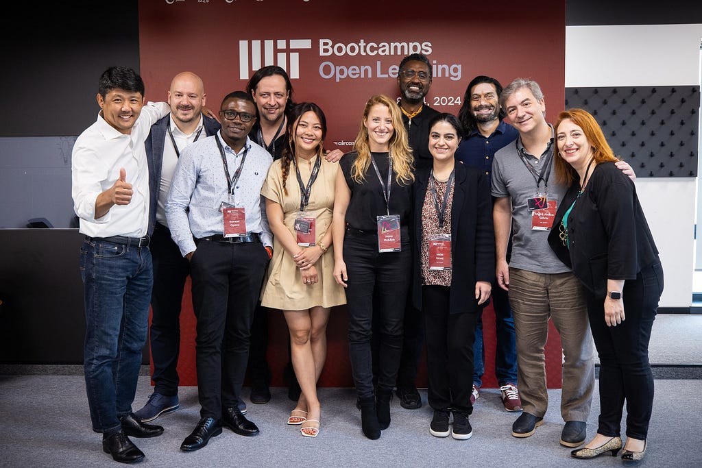 Group photo of 11 smiling adults wearing business clothes and lanyards standing in front of a sign for MIT Bootcamps at Open Learning.