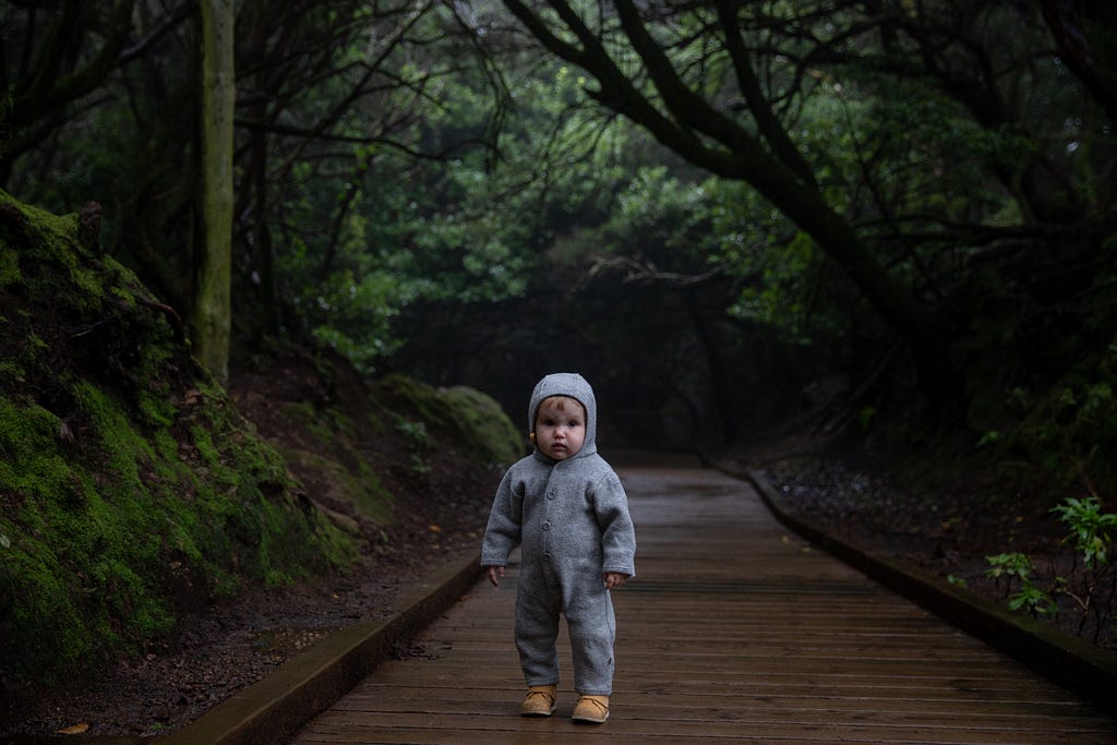 A scared-looking Infant toddler in a grey hooded onesie standing on a wood board path in the middle of a lush green forest.