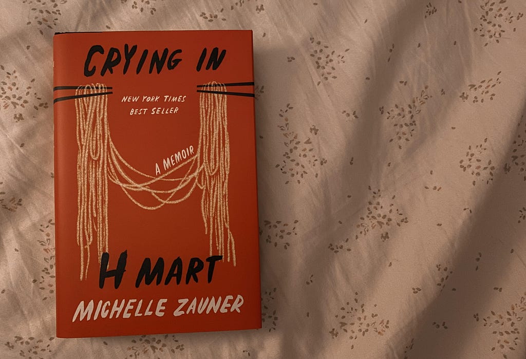 The book “Crying in H Mart” by Michelle Zauner is laying flat on a white sheet with a subtle floral pattern .