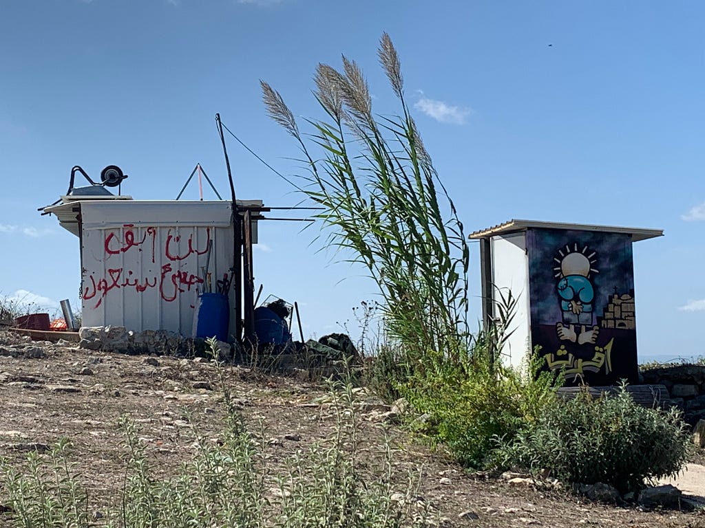 Shacks on the site of Iqrit, a Christian village in northern Palestine destroyed in 1948. The mural is of Palestinian cartoonist Naji al-Ali’s character, Handalah. The writing on the other shack reads, “I will not remain a refugee; I will return!” (Photo by the author, Sep. 30, 2023).